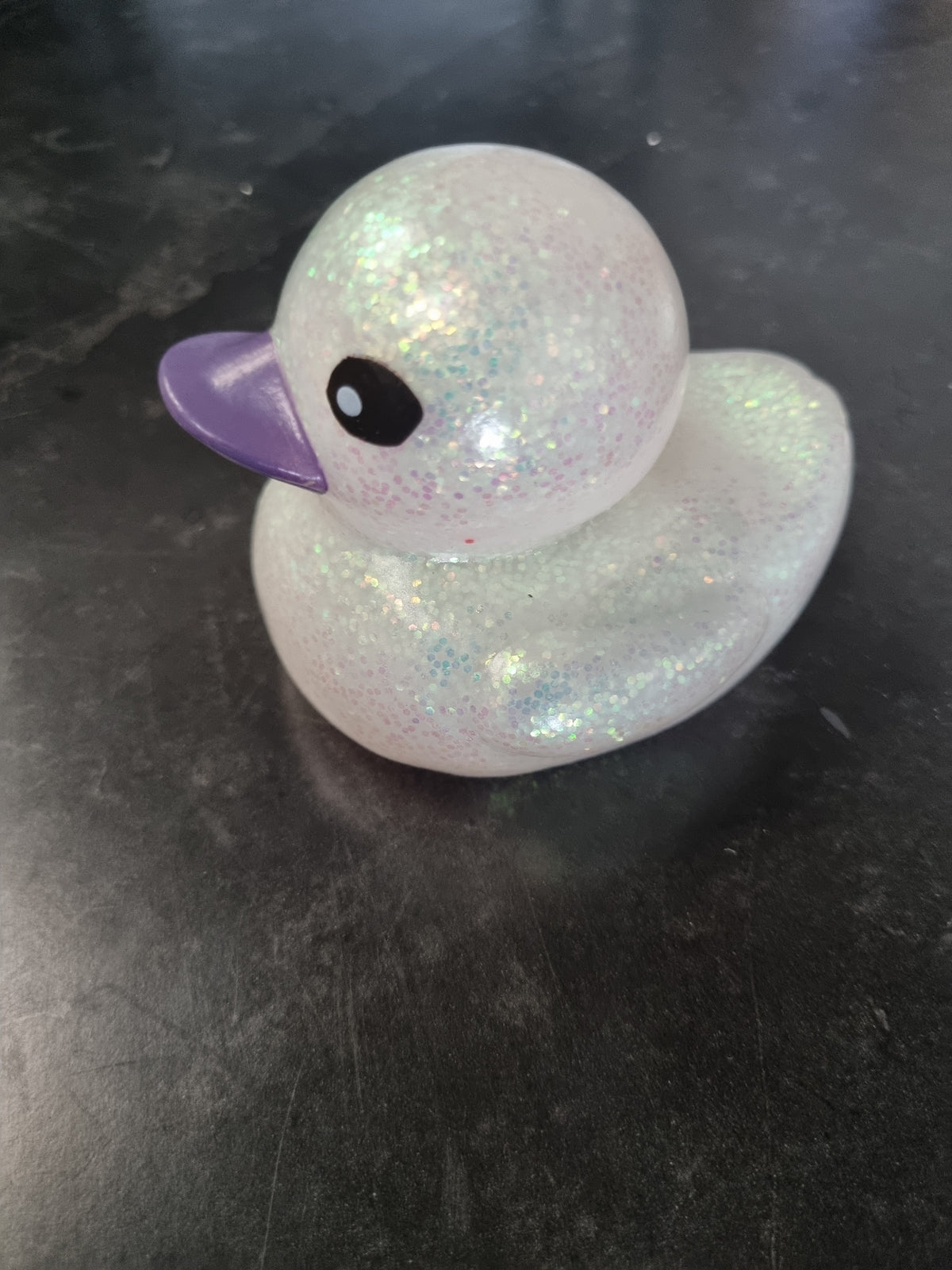 A small glittery rubber duck with a translucent white body, iridescent sparkle, and a purple beak, resting on a dark surface.