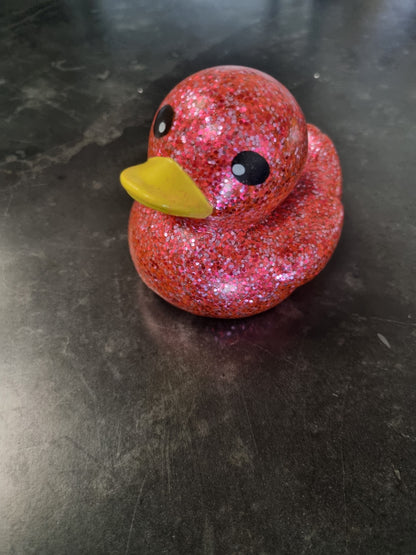 A small rubber duck toy covered in sparkling pink glitter, featuring a bright yellow beak and black painted eyes, sitting on a dark gray surface.