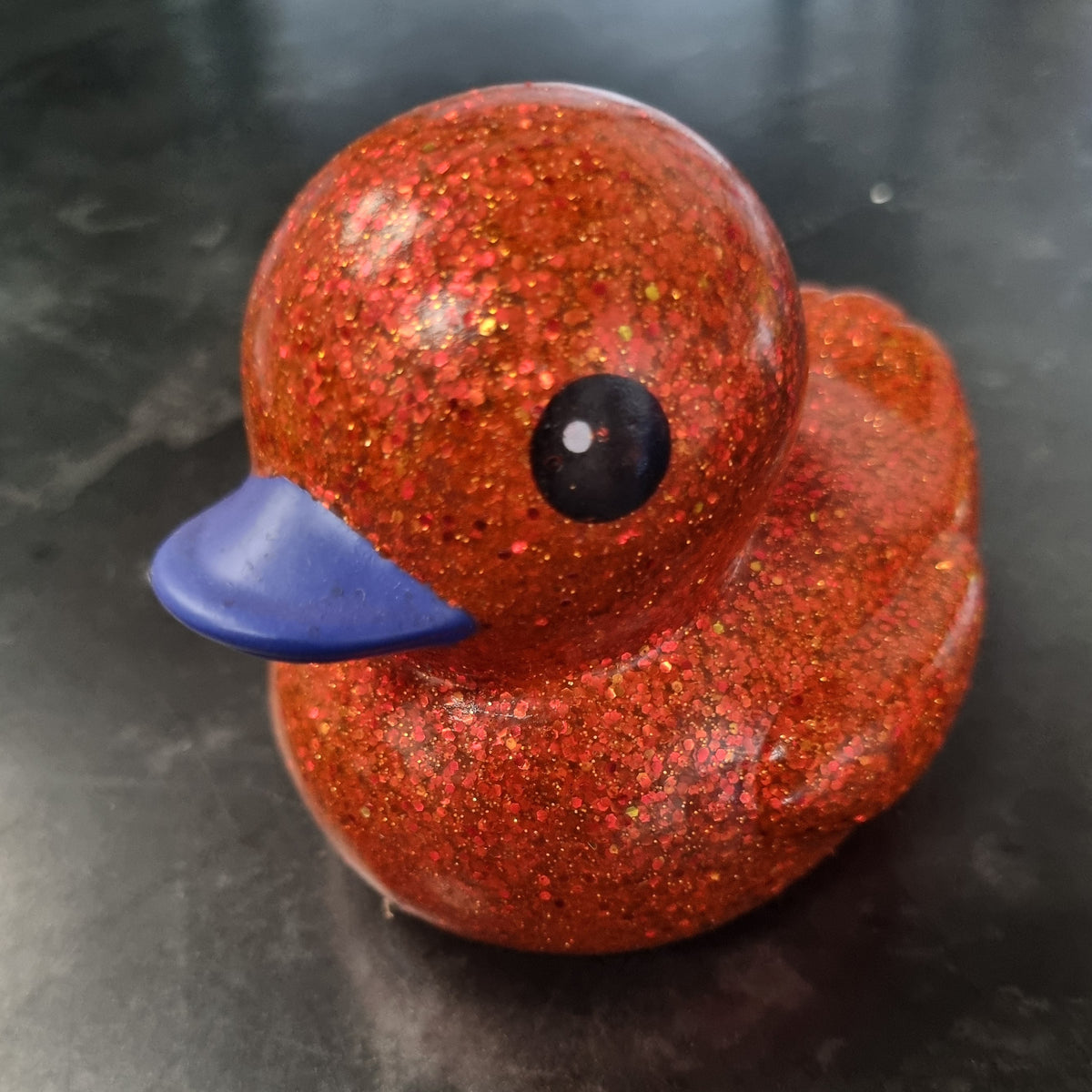 A small rubber duck toy covered in sparkling orange-red glitter, with a blue beak and a glossy black eye, sitting on a dark gray surface.