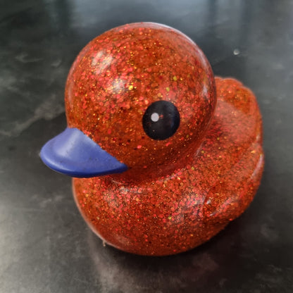 A small rubber duck toy covered in sparkling orange-red glitter, with a blue beak and a glossy black eye, sitting on a dark gray surface.