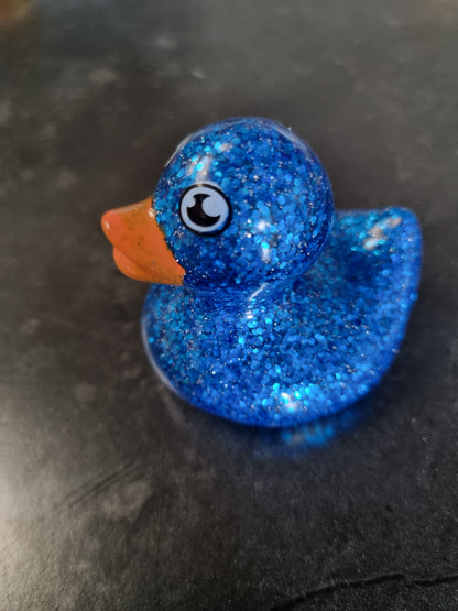 A small blue glitter rubber duck with an orange beak and a black-and-white eye, resting on a dark textured surface.