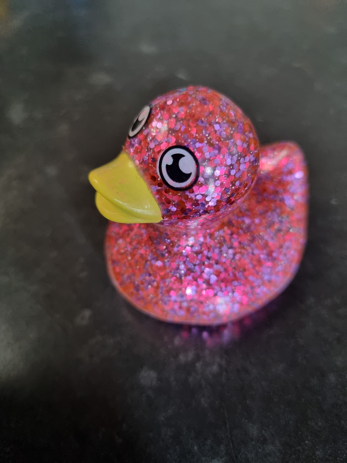 A small glittery rubber duck toy with pink and red sparkle, a yellow beak, and cartoon eyes, resting on a dark surface.