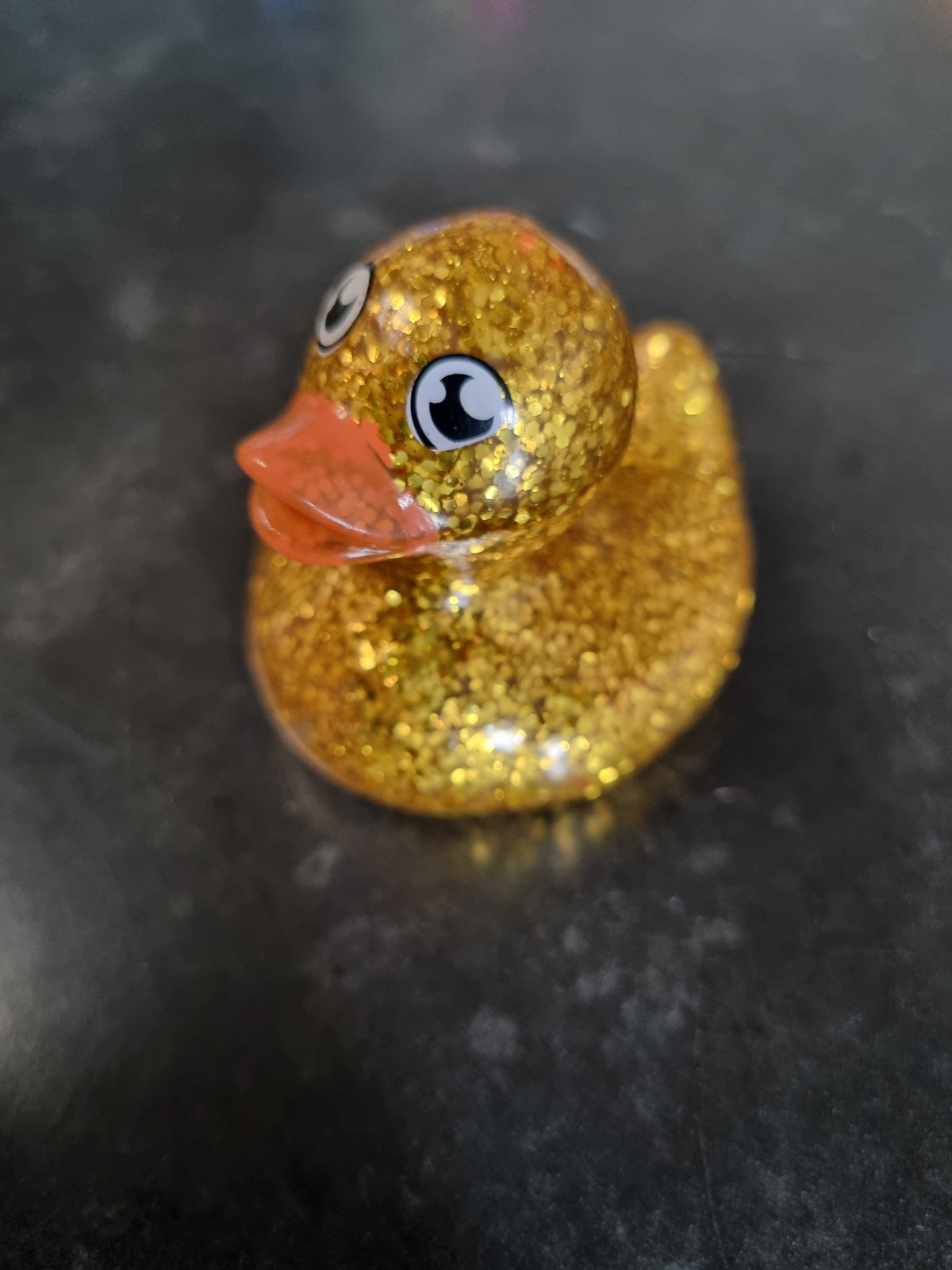 A small rubber duck toy covered in sparkling gold glitter, featuring a translucent orange beak and black-and-white cartoon eyes, resting on a dark gray surface.