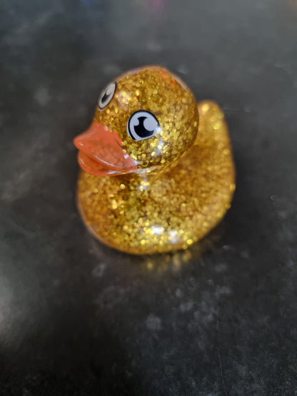 A small rubber duck toy covered in sparkling gold glitter, featuring a translucent orange beak and black-and-white cartoon eyes, resting on a dark gray surface.
