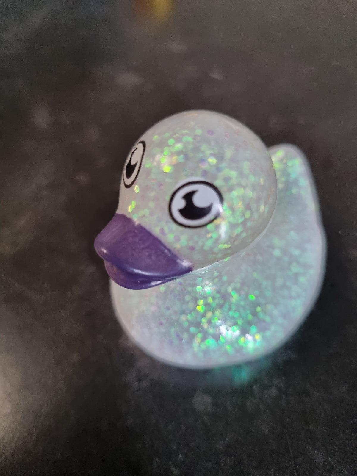 A small translucent rubber duck toy with iridescent glitter and a purple beak, photographed on a dark surface.