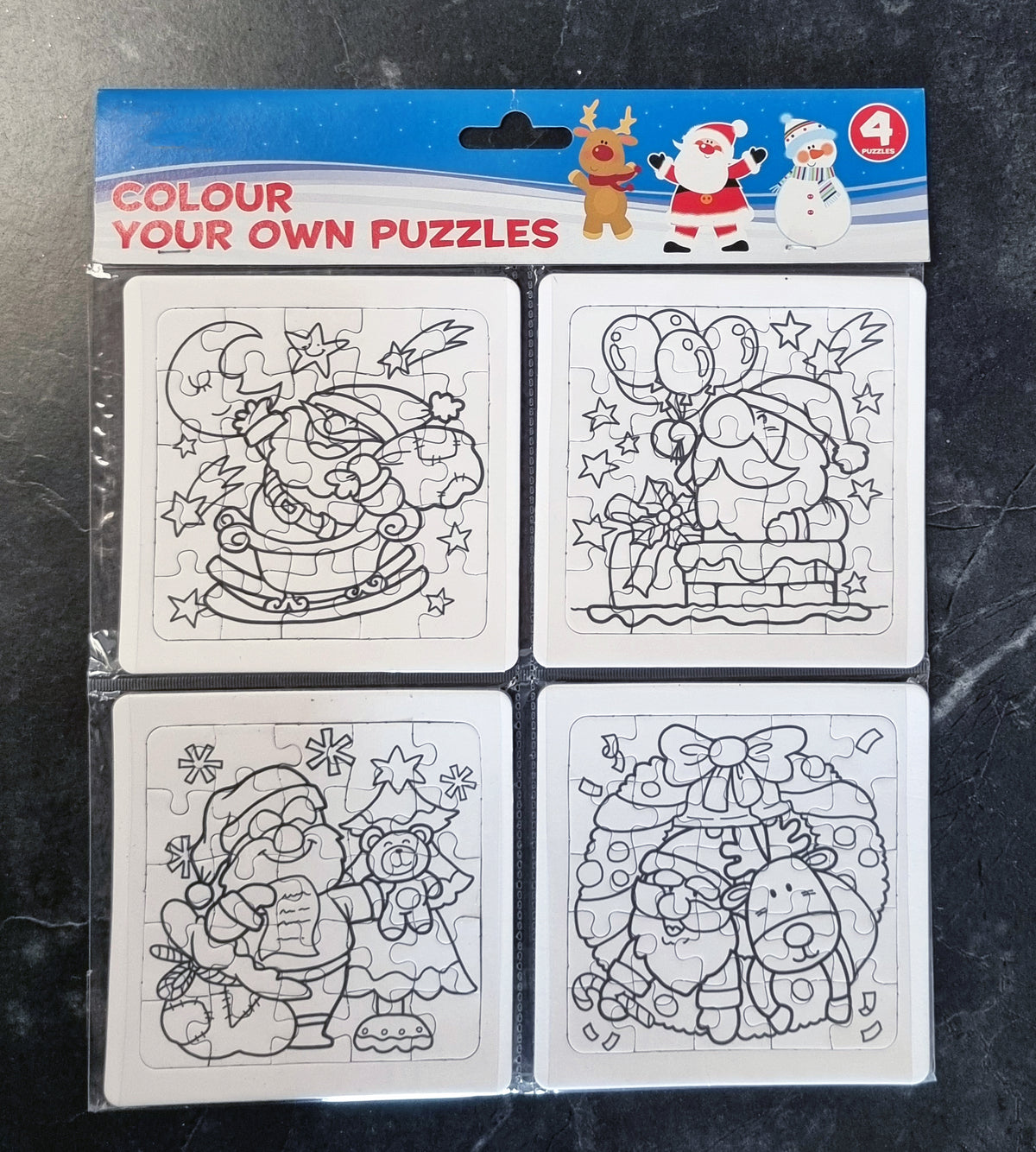 A package of four Christmas-themed colour your own puzzles on a dark marble surface. The packaging header is blue and red, and the four white puzzle cards feature black-and-white line drawings of Santa Claus, a reindeer, and a snowman.