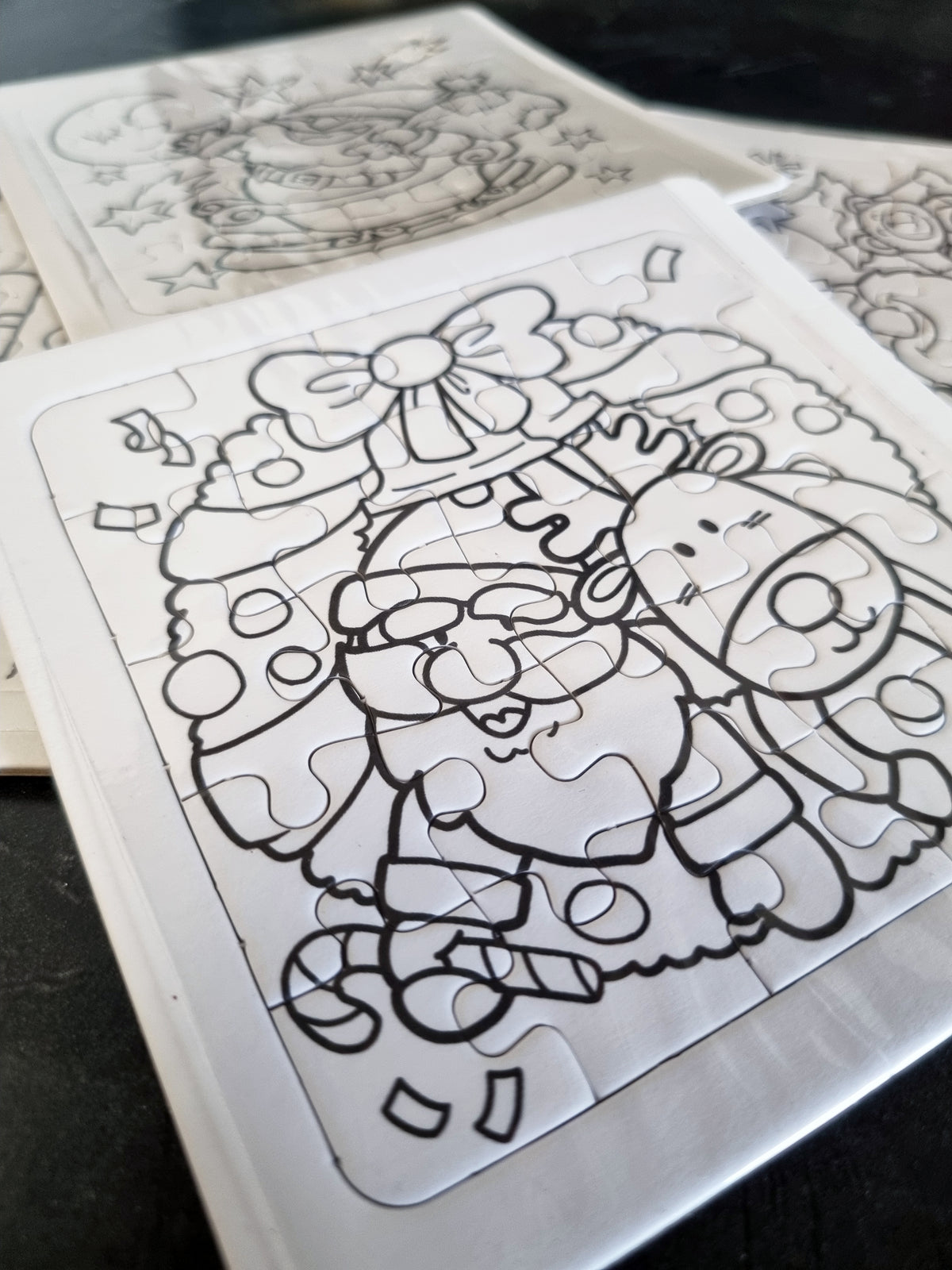 A close-up of a completed jigsaw puzzle featuring a black-and-white Christmas illustration with Santa Claus, a reindeer, and a decorated tree, placed on a dark surface with other partially visible puzzles behind it.