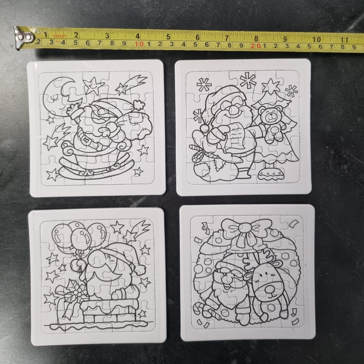 Four uncompleted black and white Christmas-themed jigsaw puzzles arranged in a 2x2 grid on a dark surface, with a yellow tape measure above them for scale.