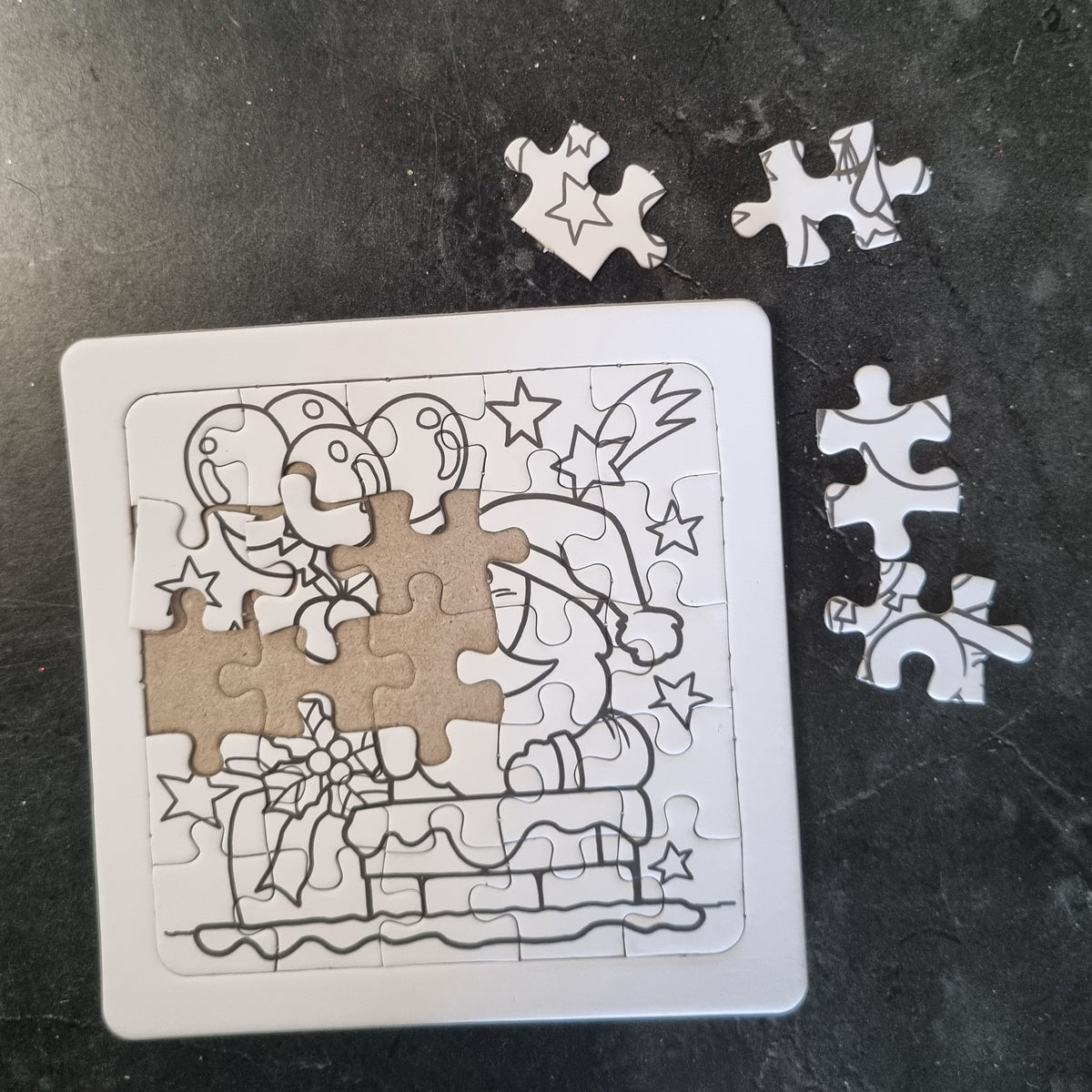 A square jigsaw puzzle with a black-and-white line drawing of a Santa Claus figure and gift, partially assembled on a dark textured surface, with several loose white puzzle pieces around it.