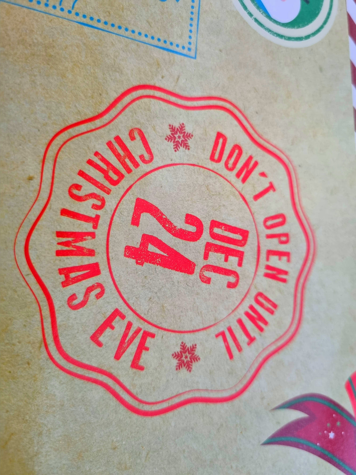 A close-up of a Christmas box surface with a red scalloped stamp reading 'CHRISTMAS EVE DON'T OPEN UNTIL 24 DEC' and small snowflake details.