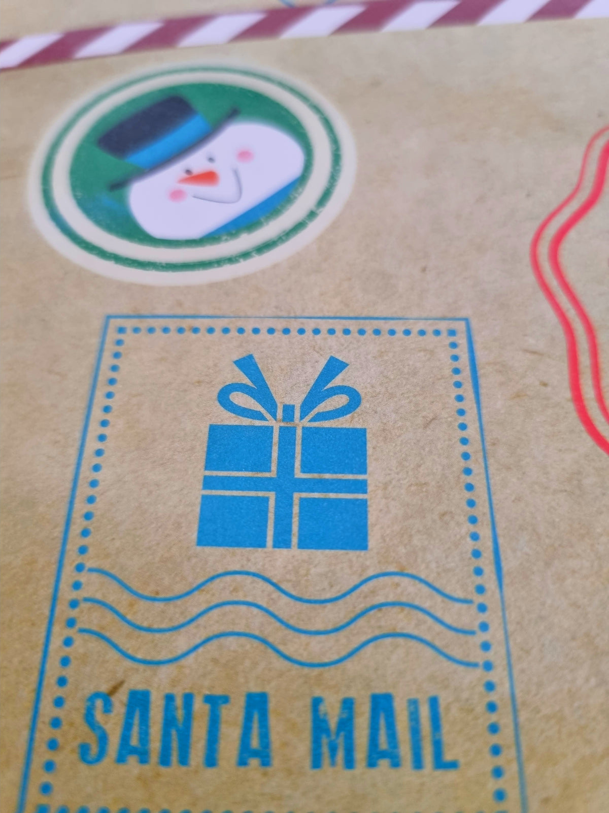 A close-up of a kraft paper envelope or card featuring a blue 'SANTA MAIL' gift box stamp design and a circular snowman sticker.