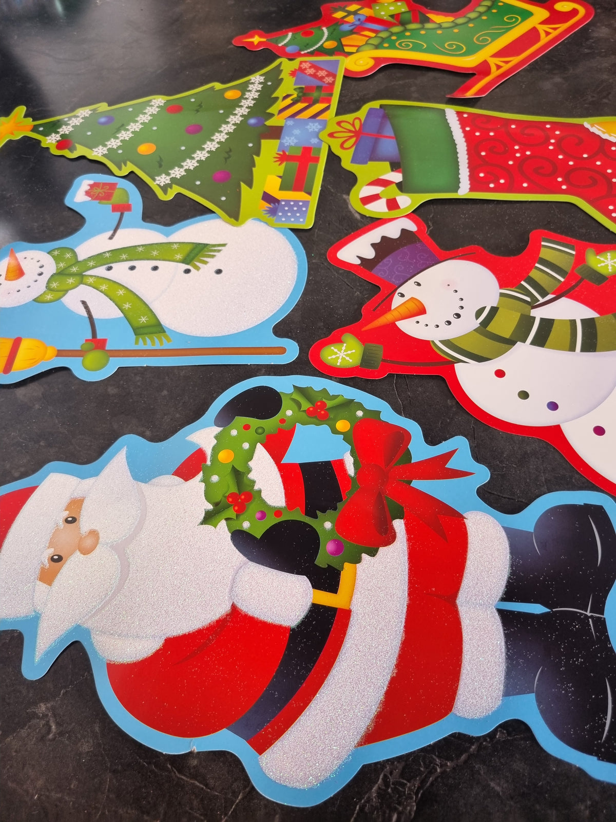 A set of colorful Christmas-themed felt cutouts arranged on a dark marble surface, featuring Santa Claus, snowmen, stockings, a Christmas tree, and a gift box.