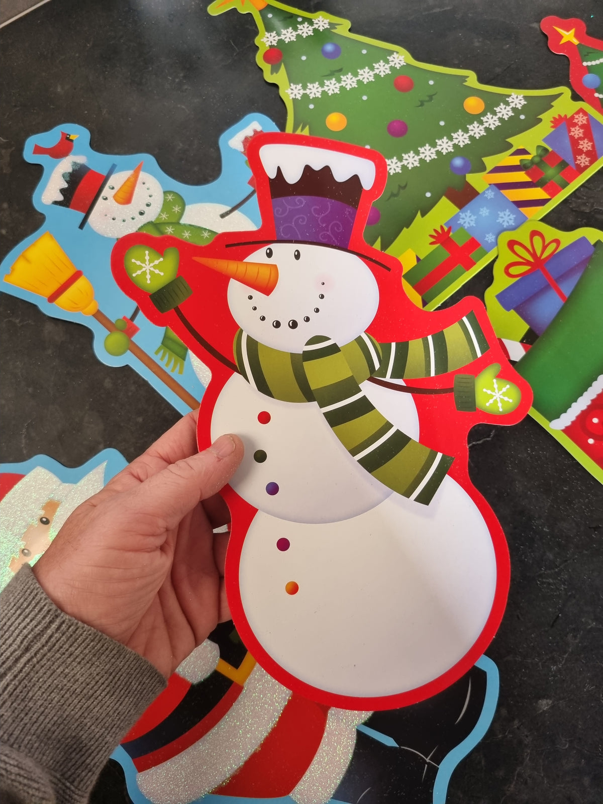 A hand holding a colorful Christmas snowman craft cutout, with other holiday-themed paper cutouts like a Christmas tree and gifts visible in the background on a dark surface.