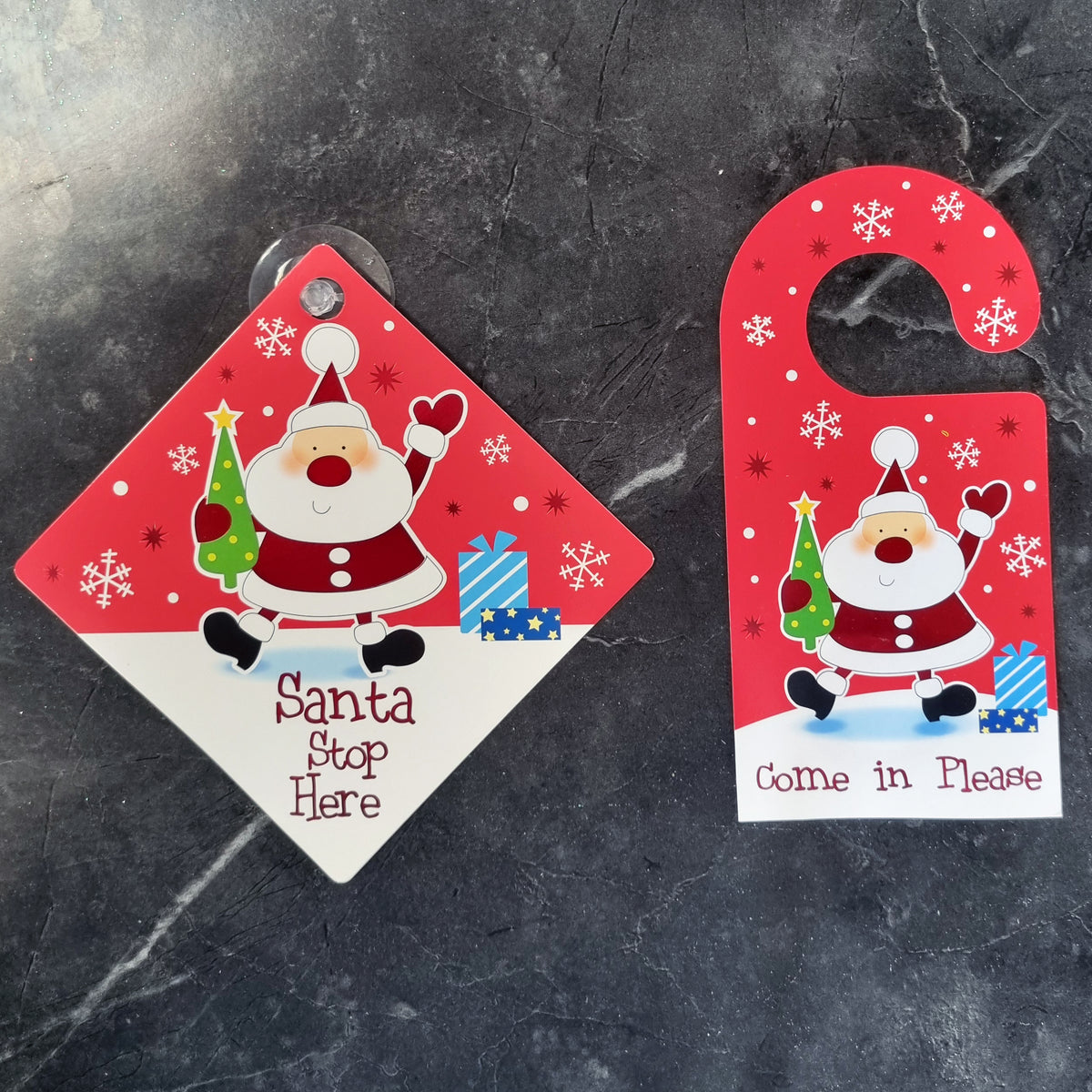 Two Christmas-themed door signs on a dark gray marble surface: a diamond-shaped sign that reads 'Santa Stop Here' with a Santa illustration, and a red hanger-style door hanger sign that reads 'Come in Please' with the same Santa design.