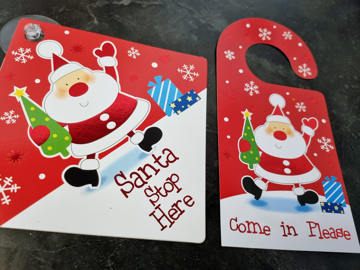 Two red and white Christmas-themed door hangers on a dark textured surface. One says 'Santa Stop Here' and the other says 'Come in Please', both featuring a cartoon Santa holding a tree and blue wrapped gifts.