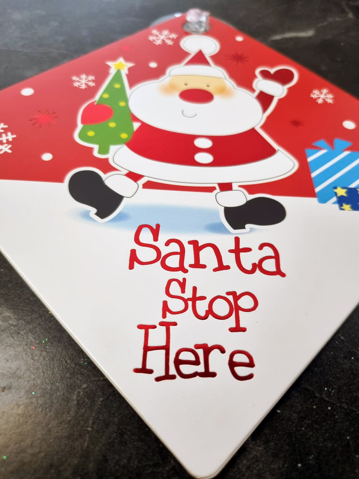 A diamond-shaped Christmas sign featuring a cartoon Santa Claus and a small tree on a red background, with the words 'Santa Stop Here' in red script on a white lower section, photographed on a dark surface.