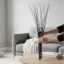Load image into Gallery viewer, Person arranging branches in a clear vase on a table with a gray sofa and books in the background.
