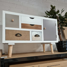 Load image into Gallery viewer, A mid-century modern storage cabinet with a white frame, mixed wood drawer fronts, burlap texture panels, and light wood legs, sitting on a shelf in a warehouse setting.
