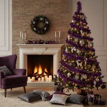 Load image into Gallery viewer, A decorated Christmas tree in a cozy living room beside a lit fireplace. The tree is wrapped in metallic purple tinsel with warm white string lights, silver and white ornaments, reindeer figures, and bows, with wrapped gifts underneath.
