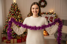 Load image into Gallery viewer, A woman in a cream knit sweater holds up a bright purple metallic tinsel garland in a cozy, decorated living room with a Christmas tree, fireplace, and stockings in the background.
