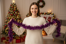 Load image into Gallery viewer, A woman in a cream knit sweater holds up a bright purple metallic tinsel garland in front of a cozy Christmas living room background with a decorated tree, fireplace, and stockings.
