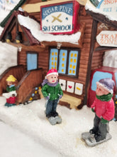 Load image into Gallery viewer, A miniature winter village scene featuring a rustic log cabin labeled 'Sugar Pine Ski School' with snow on the roof, small figurines of children in winter clothing standing in the snow, and a red vehicle on the right.
