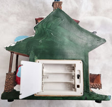 Load image into Gallery viewer, A small painted house-shaped object with a green roof and side details, shown with its front door open to reveal battery compartments inside. The item appears to be the product itself rather than packaging, photographed against a light fabric background.
