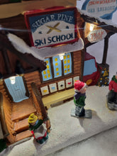 Load image into Gallery viewer, A miniature Christmas village scene featuring a wooden cabin labeled 'Sugar Pine Ski School' with small figurines of children in winter clothing standing outside on a snowy ground.
