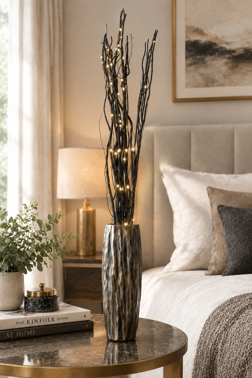 A tall black branch-style vase with warm white string lights, placed on a round marble nightstand in a softly lit bedroom. The scene includes a bed, lamp, books, and framed wall art.