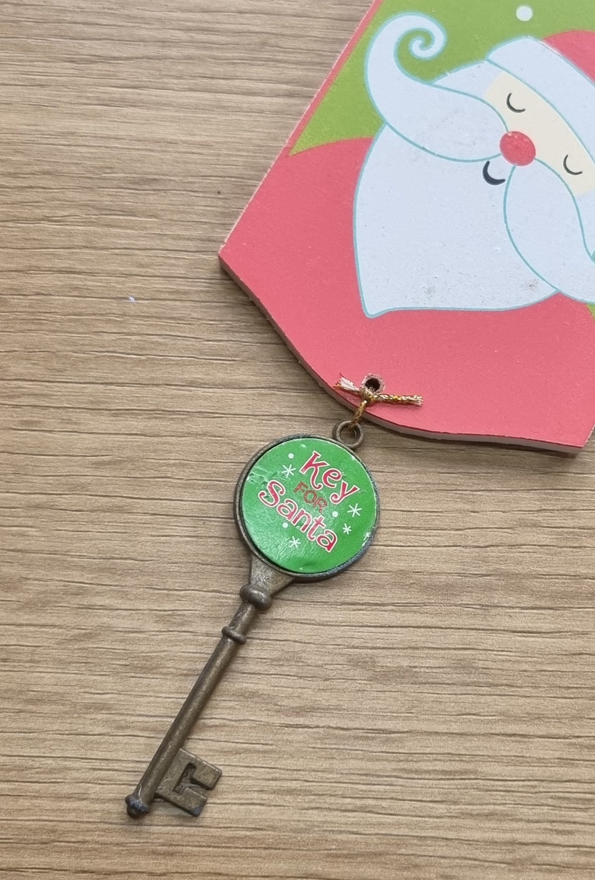 A vintage-style metal key with a round green pendant engraved with 'Key for Santa' in red and white, resting on a light wood surface next to a Christmas-themed card featuring Santa's face.