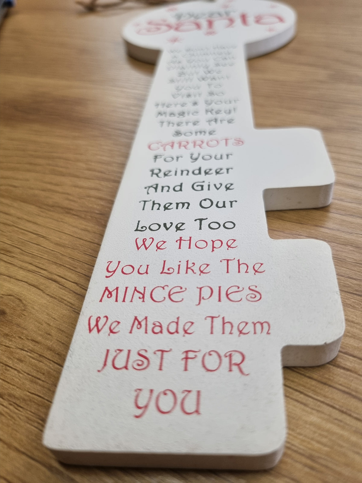 A white wooden Santa-shaped Christmas message card on a wooden tabletop, with red and black printed text and a second similar card partially visible in the background.
