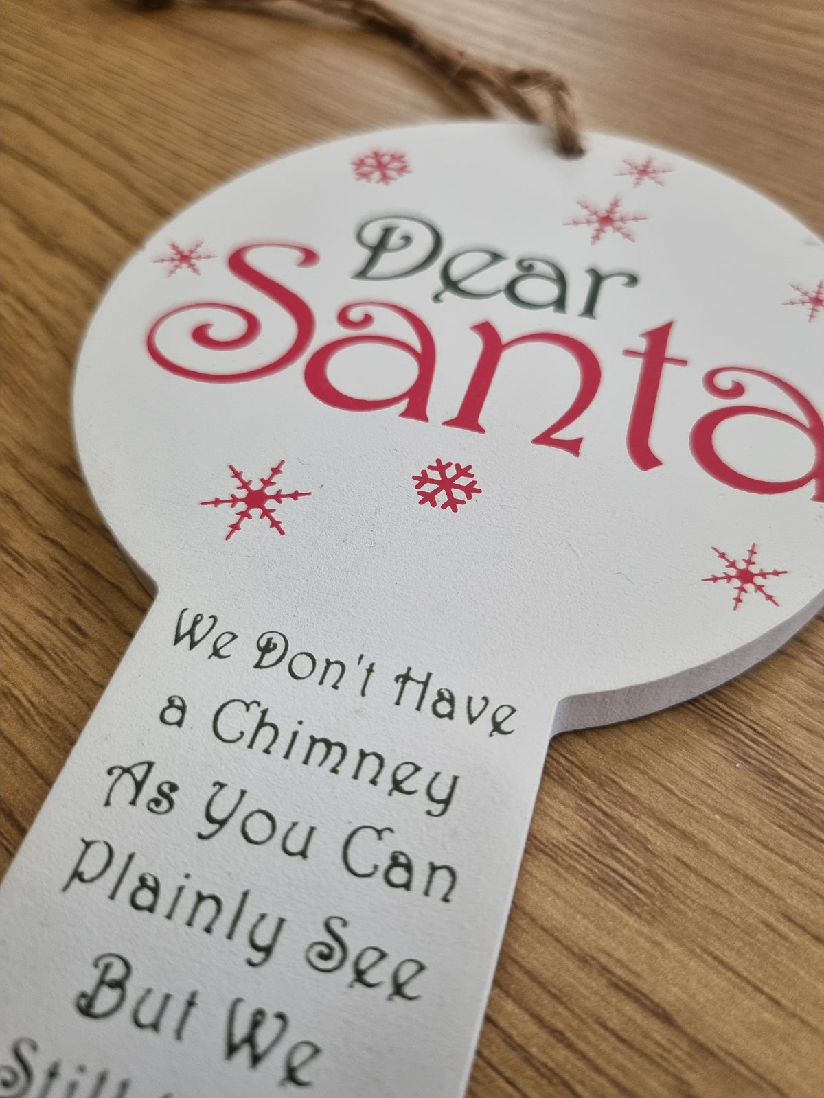 A close-up of a round white Christmas ornament or tag with a hanging string, printed with the words 'Dear Santa' in dark green and red script, red snowflake motifs, and a handwritten message on a wooden surface.