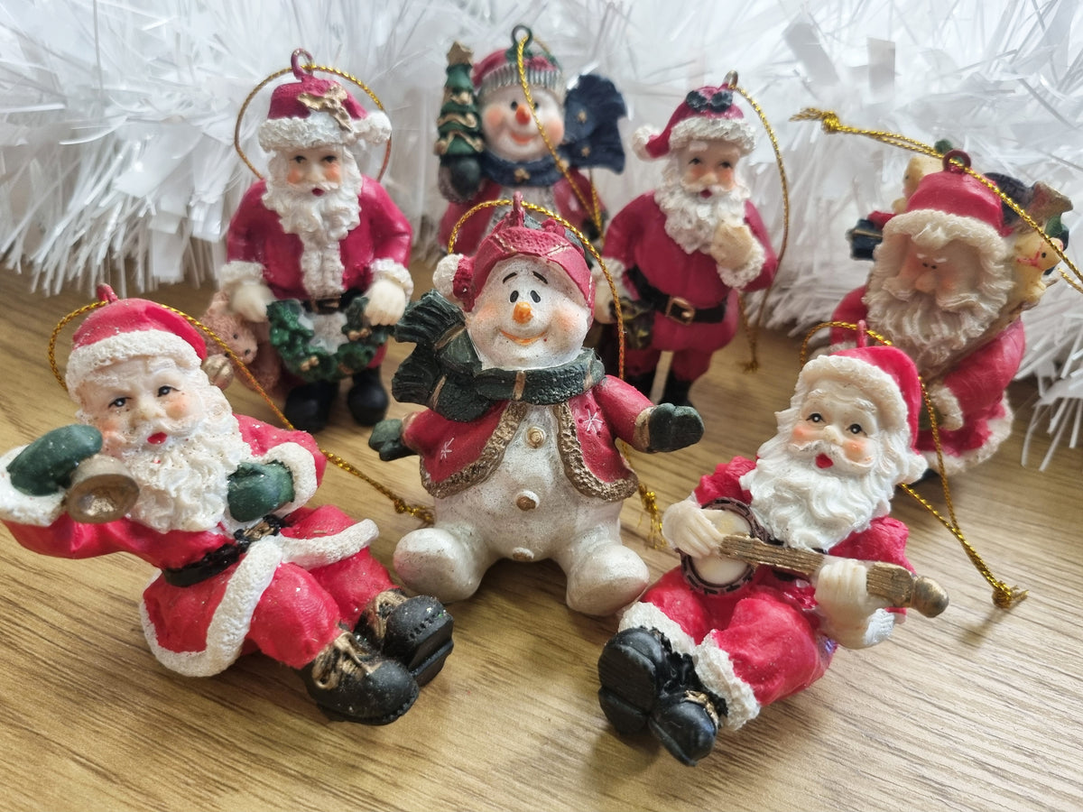 A group of Christmas ornaments shaped like Santa Claus and a snowman, displayed on a wooden surface with white tinsel in the background.