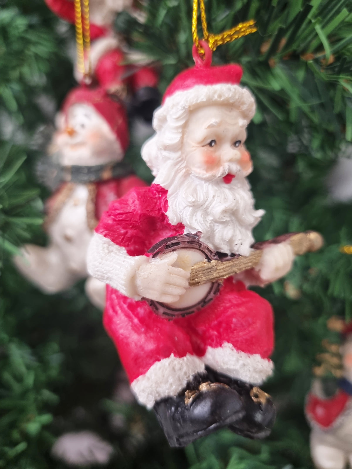 A close-up of a Santa Claus Christmas ornament playing a banjo, hanging on a green Christmas tree. The ornament features red and white clothing with black boots and a gold hanging string.