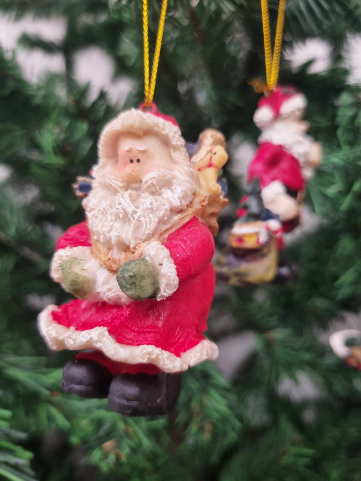 A Santa Claus hanging ornament with a red suit, white beard, and black boots, suspended by a yellow string on a green Christmas tree. Another ornament is blurred in the background.