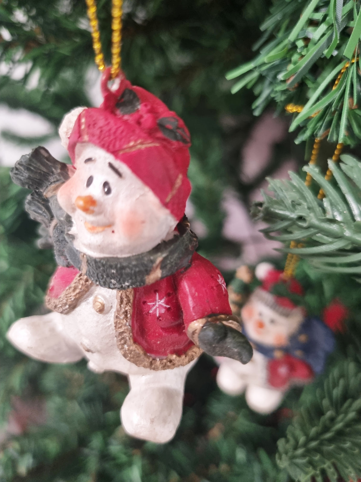 A close-up of a small snowman Christmas ornament hanging from a green Christmas tree. The ornament is dressed in a red coat and hat with a dark gray scarf, and another similar ornament is blurred in the background.