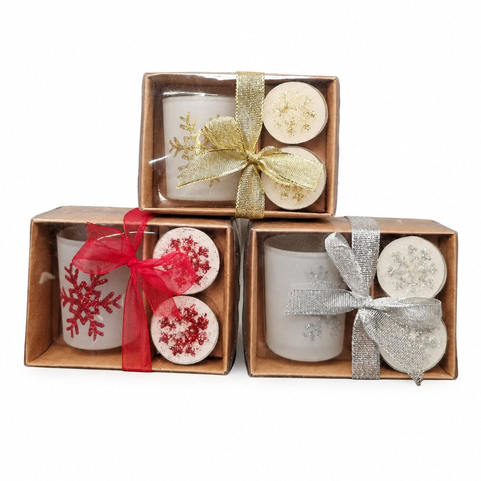 Three small gift boxes containing candles arranged on a white background. The box has white candles with a gold ribbon, the bottom-right box has white candles with red snowflake designs and a red ribbon, and the bottom box has white candles with a red ribbon.