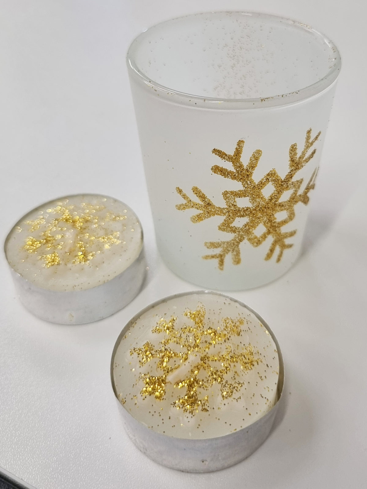 A frosted glass tea light holder with gold glitter decorated on the front with 2 tea light candles also decorated with gold glitter sitting next to it on a white table