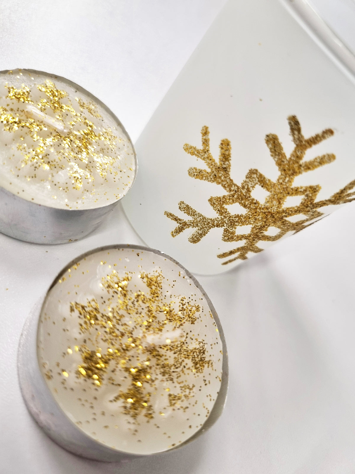 Two round white candles in silver metal cups, each topped with scattered gold glitter, next to a white cylindrical container with a gold glitter snowflake design on a white background.