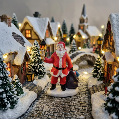 A festive Christmas mini village scene with a Santa Claus figurine standing on a cobblestone path between snow-covered houses, lit by warm yellow window lights and lanterns.
