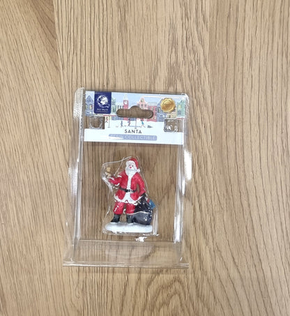 A Santa Claus miniature figurine in its clear plastic blister packaging, displayed on a light wood grain surface.