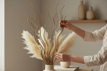 Load image into Gallery viewer, Person arranging dried pampas grass and branches in a white vase on a wooden surface.
