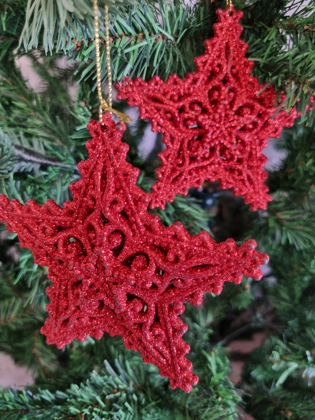 Two red glittery star-shaped Christmas ornaments hanging on a green Christmas tree branch.