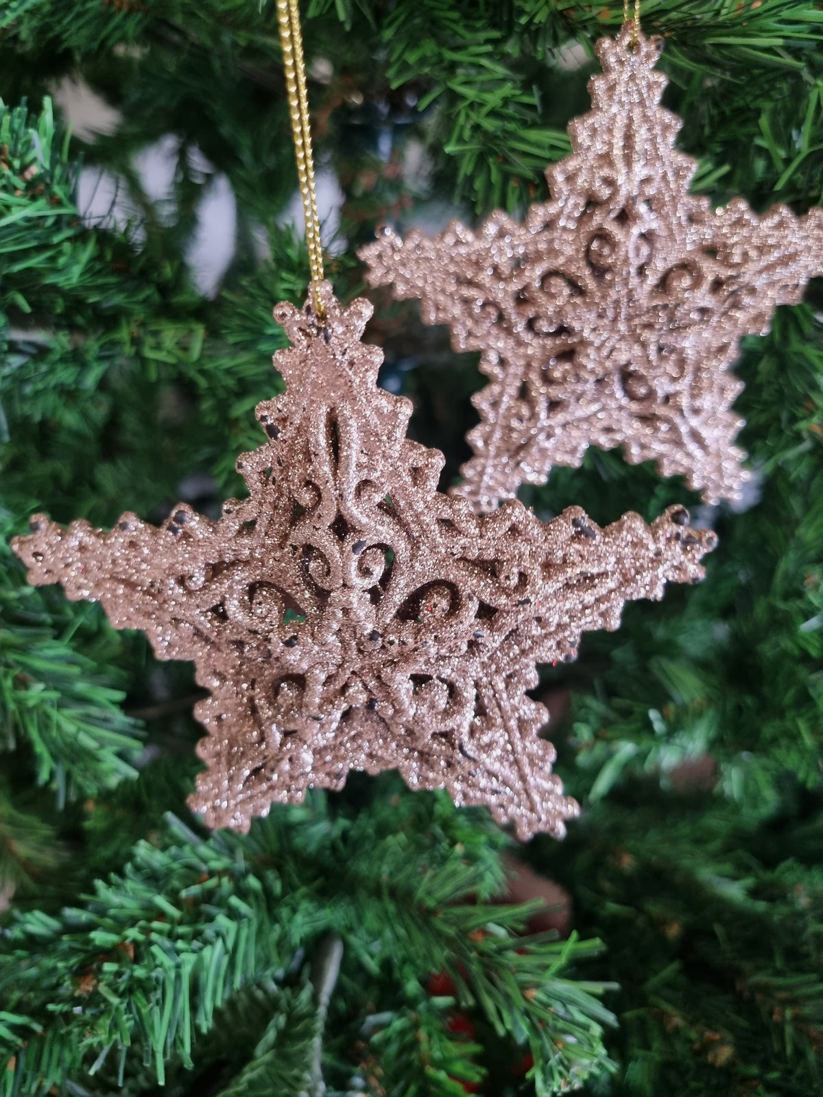 Two glittery rose gold star-shaped Christmas ornaments hanging from a gold string on a green Christmas tree.