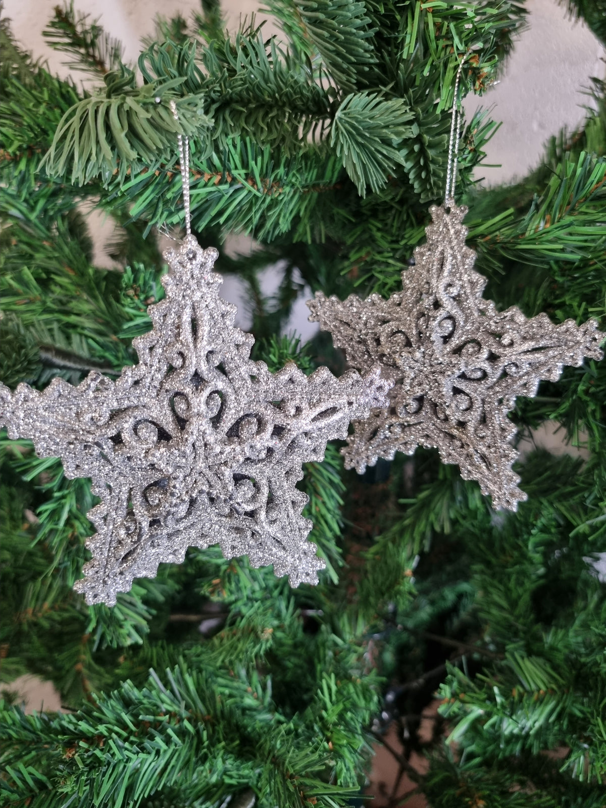 Two glittery silver snowflake Christmas tree ornaments hanging from green artificial pine branches.