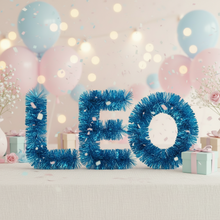 Load image into Gallery viewer, Three blue tinsel letters spelling LEO are displayed on a table with pastel balloons, confetti, and gift boxes in the background.
