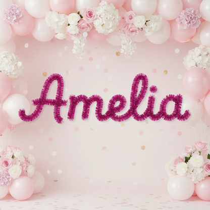 Personalized party backdrop with the name 'Amelia' spelled in large magenta tinsel letters, surrounded by pastel pink and white balloons, white and pink flowers, and scattered confetti on a light pink background.
