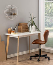 Load image into Gallery viewer, A modern white desk with light wood legs, styled in a home office setting next to a brown leather swivel chair, with decorative items on top.
