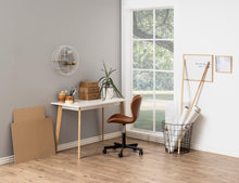 Load image into Gallery viewer, A styled home office corner featuring a white desk with light wood legs, a brown leather swivel office chair, a wire wall shelf, and a window looking out to green trees. Decorative items like a plant, cardboard boxes, and art supplies are arranged on the floor and desk.
