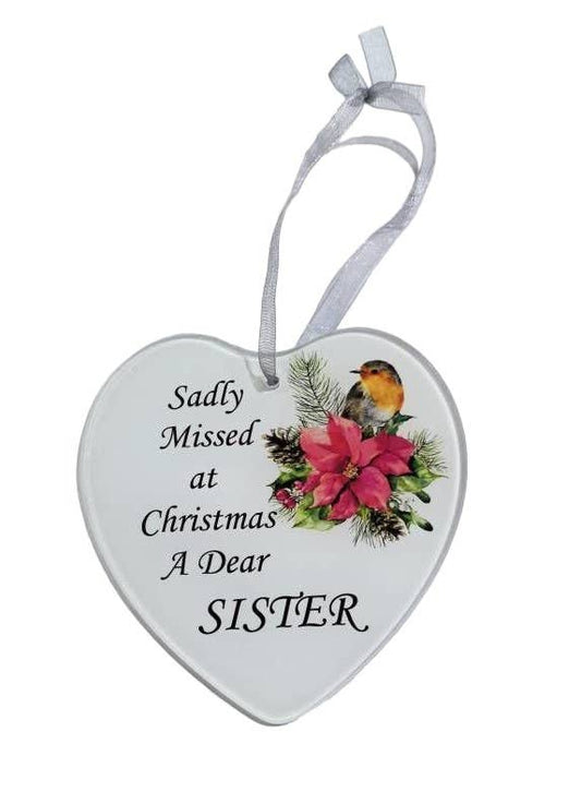 A white heart-shaped glass hanging decoration with a message 'Sadly missed at Christmas a dear Sister' and an image of a robin and poinsettia flowers.